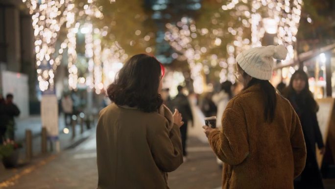 两个亚洲女人的后视图，在许多圣诞灯的夜晚在城市里散步和聊天。