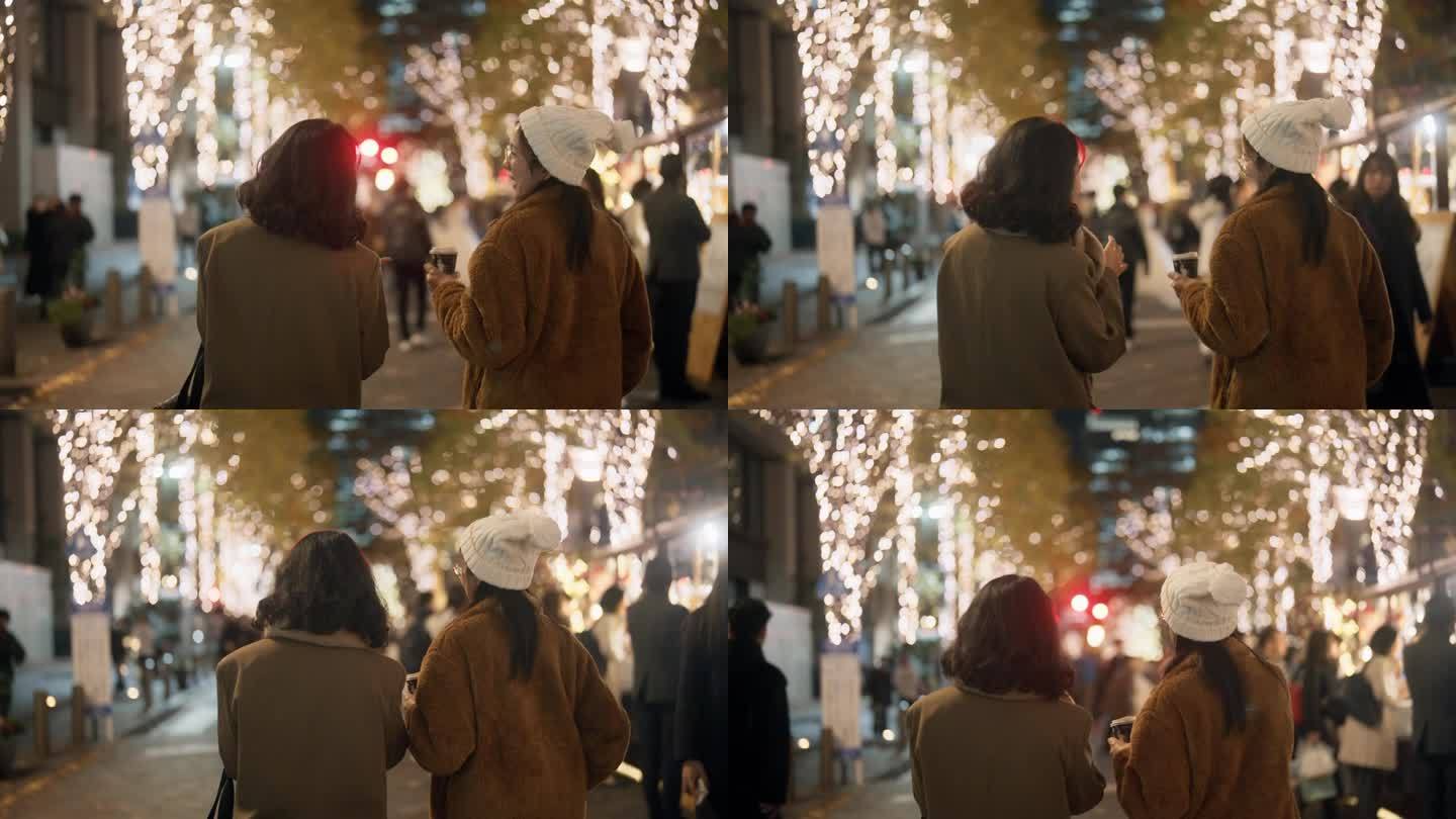 两个亚洲女人的后视图，在许多圣诞灯的夜晚在城市里散步和聊天。