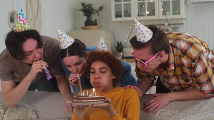 许个愿。戴着派对帽的女子吹灭了生日蛋糕上燃烧的蜡烛。生日派对快乐。朋友群祝女孩生日快乐。人们在家里开