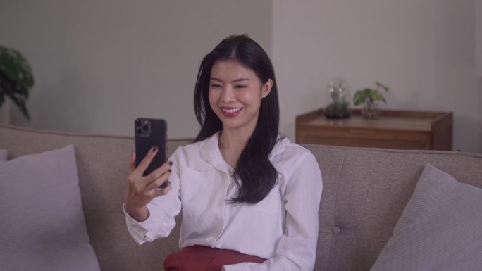微笑的年轻迷人的亚洲女人，用智能手机，在社交网络上挥手，在家里的沙发上聊天。