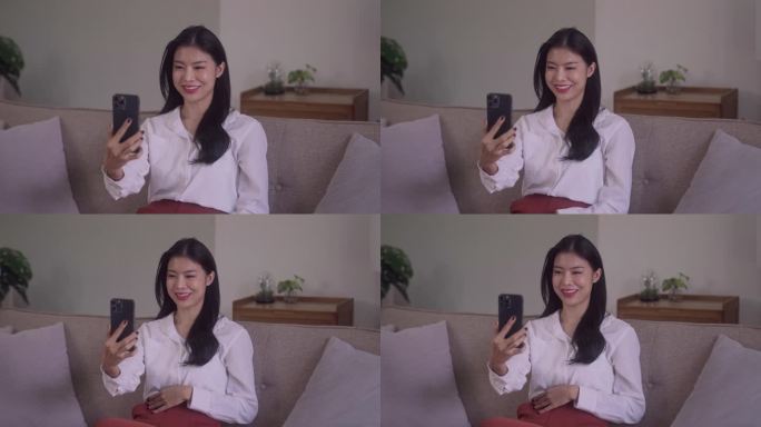 微笑的年轻迷人的亚洲女人，用智能手机，在社交网络上挥手，在家里的沙发上聊天。