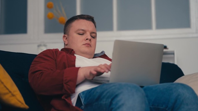 胖胖的年轻人坐在沙发上用笔记本电脑工作，不健康的久坐生活