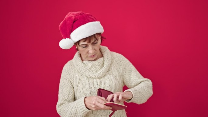 成熟的西班牙女人戴着圣诞帽，看着孤立的红色背景上空空的钱包