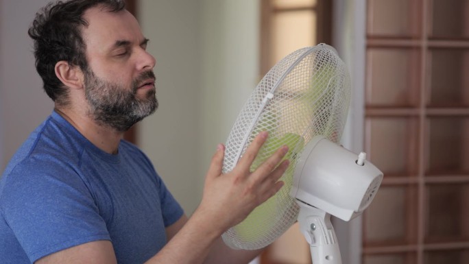 夏日男子在家享受电扇吹来的凉风。一名男子在通风机前酷暑，用电扇冷却器降温。炎热的天气，气候变化。