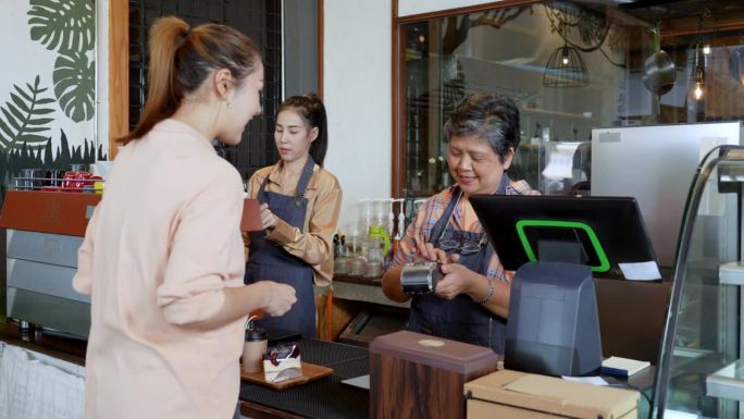 漂亮的亚洲女性顾客在面包店和咖啡店买蛋糕。围裙上的老店主卖主正在安排付款，而年轻的女士正在摆弄咖啡机