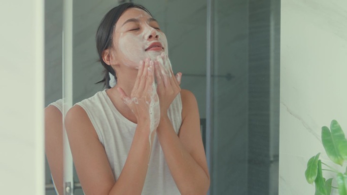 上午晚些时候，年轻的亚洲女人在家里的厕所里对着镜子用泡沫洗脸。日常的生活方式。