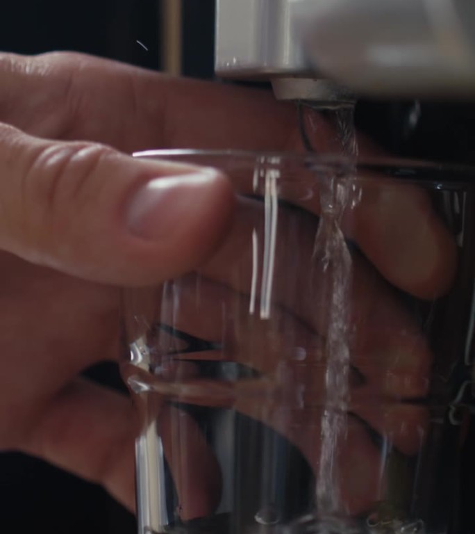 男性手从饮水机向玻璃杯中倒入淡水的垂直方向