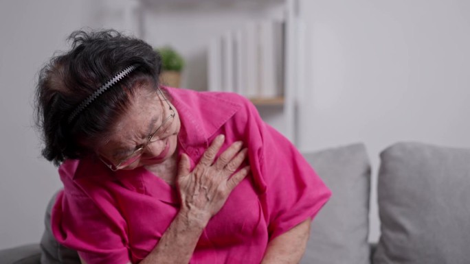 一位患有心脏病的亚洲老年祖母坐在客厅的沙发上捂着胸口。医疗急救和医疗保险保障理念