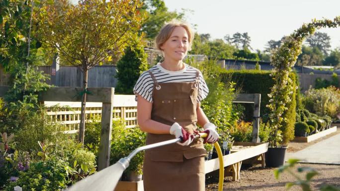 一名身穿围裙的妇女在花园中心的户外用软管给植物浇水——慢镜头