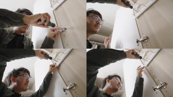 一个男人在家里DIY平板家具组装时，用螺丝刀打开壁橱门，拧紧铰链上的螺丝，这是一个低角度向上倾斜的镜