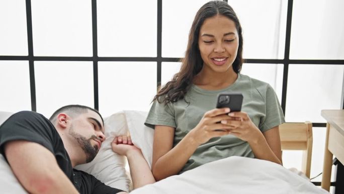 美丽的情侣惬意地躺在床上，女友玩智能手机，而打盹的男友照亮了房间，一起享受他们安静的室内早晨