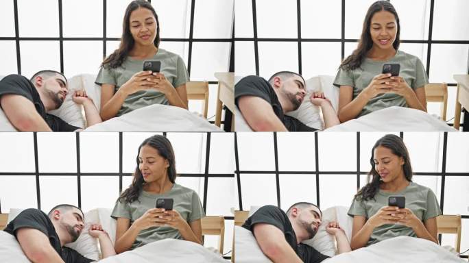 美丽的情侣惬意地躺在床上，女友玩智能手机，而打盹的男友照亮了房间，一起享受他们安静的室内早晨