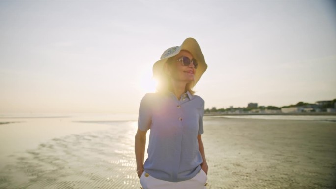 一位戴着帽子和太阳镜的老年妇女在日落时分走在海滩上