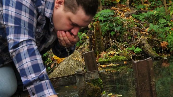 一个男人弯下腰，在泉水旁的一条干净的森林河流里喝水。人用手打水，在源头止渴。纯净清凉的泉水恢复体力。
