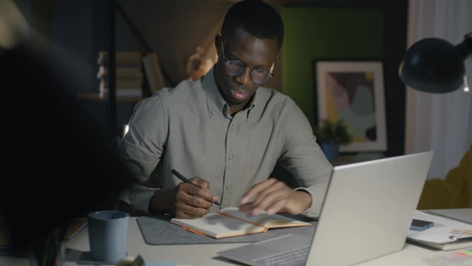非裔美国商人晚上在办公室使用笔记本电脑并在笔记本上记录信息