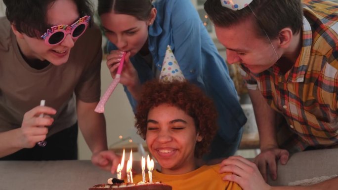 许个愿。戴着派对帽的女子吹灭了生日蛋糕上燃烧的蜡烛。生日派对快乐。朋友群祝女孩生日快乐。人们在家里开