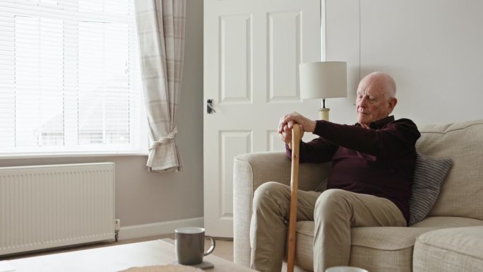老人，拄着拐杖，在家里的沙发上思考，在照顾者的支持下或孤独地退休。老年人、痴呆症患者和有残疾的老年人