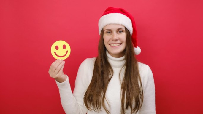 快乐有趣的年轻女子穿着白色毛衣，戴着圣诞老人帽，拿着小黄脸表情符号，在工作室的红色背景墙上独自摆姿势