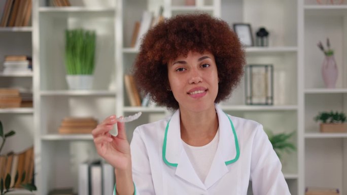女性正畸医生坐在医疗柜前，展示牙齿矫正的清晰对准器。穿着实验室大褂的非裔美国专家解释使用替代牙套的好