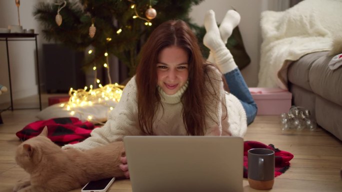快乐的黑发女孩穿着白色毛衣，躺在红色格子地毯上，在装饰成圣诞风格的舒适房间里，用笔记本电脑看电影，抚