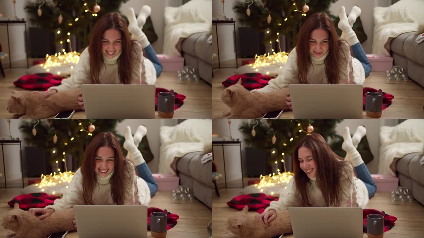 快乐的黑发女孩穿着白色毛衣，躺在红色格子地毯上，在装饰成圣诞风格的舒适房间里，用笔记本电脑看电影，抚