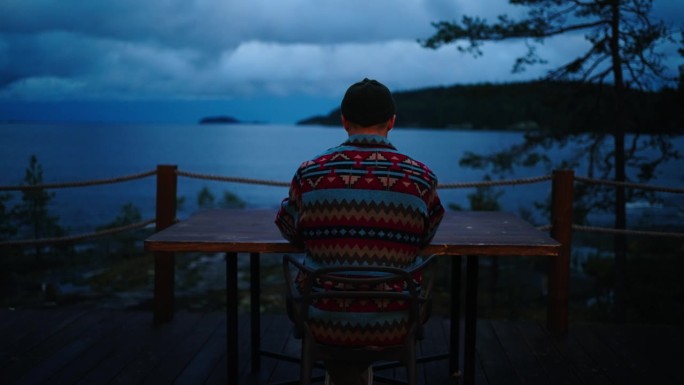 晚上，一个孤独的男人坐在阳台的桌子旁，试图用笔记本电脑工作，后景