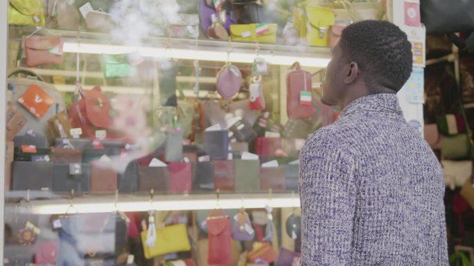 一名年轻的非洲男子站在一家皮具店的橱窗前，看着陈列的商品