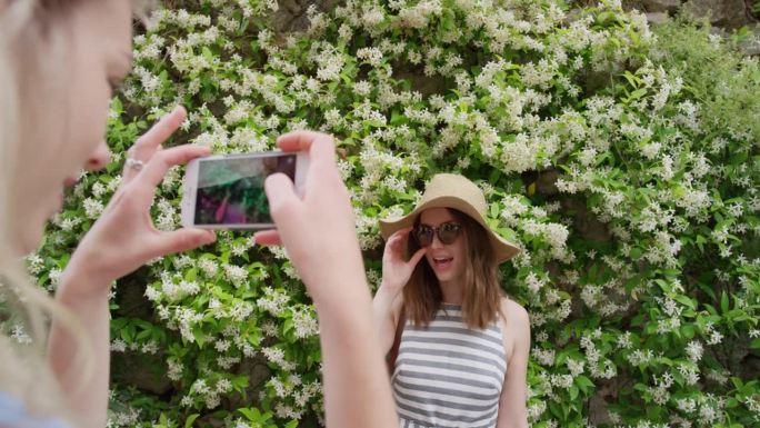 最好的朋友在度假时用智能手机自拍，头上戴着花。女孩们站在花墙前发短信，用手机分享生活方式的照片
