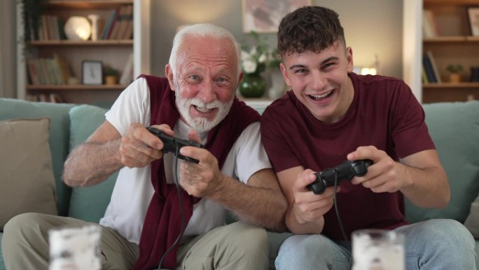 白人少年和老人爷爷和孙子坐在家里的沙发床上玩游戏机视频游戏拿着操纵杆控制器玩得很开心家庭纽带男性男性