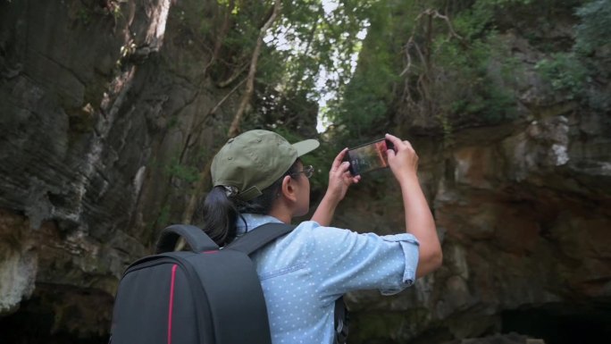 亚洲女游客正在用手机拍摄视频，记录夏天被绿色植物覆盖的岩石悬崖的美丽。