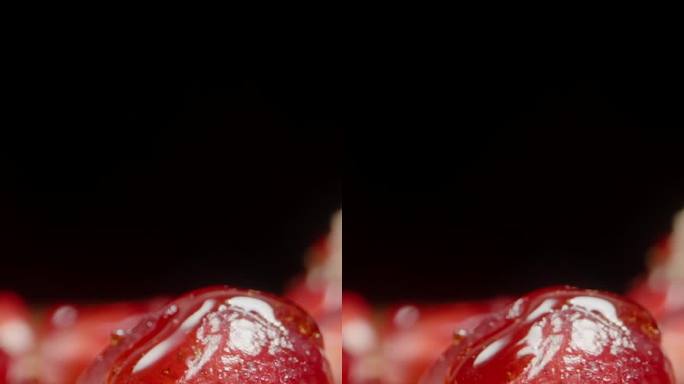 垂直视频。在黑色的背景上，红石榴籽和汁液从上面滴下来，慢慢地倾泻在种子上。特写镜头。
