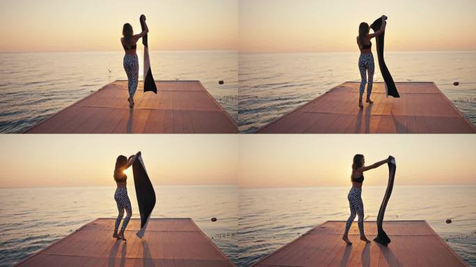 一个穿着黑白裤子和黑色上衣的年轻女孩走到码头上，在日出时在海边展开她的小运动垫做瑜伽