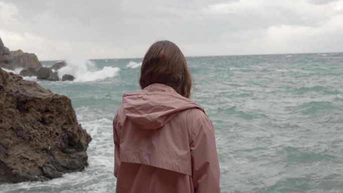 一个孤独的女人凝视着远处汹涌的大海，海浪冲击着尖锐的岩石。缓慢的运动。