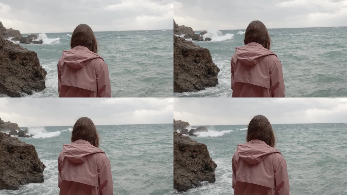 一个孤独的女人凝视着远处汹涌的大海，海浪冲击着尖锐的岩石。缓慢的运动。