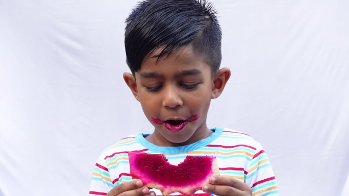 一个亚洲孩子正在吃一片粉红色的火龙果，背景是孤立的白色。一个小孩拿着或吃着新鲜的火龙果。吃新鲜水果，