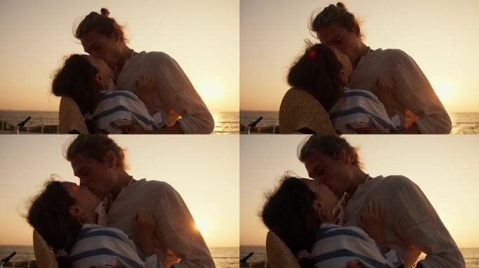 一对幸福的情侣，一个穿着浅色衣服的男人和一个穿着蓝白相间衬衫的女孩在日出时在海边接吻。夏天日出时，一