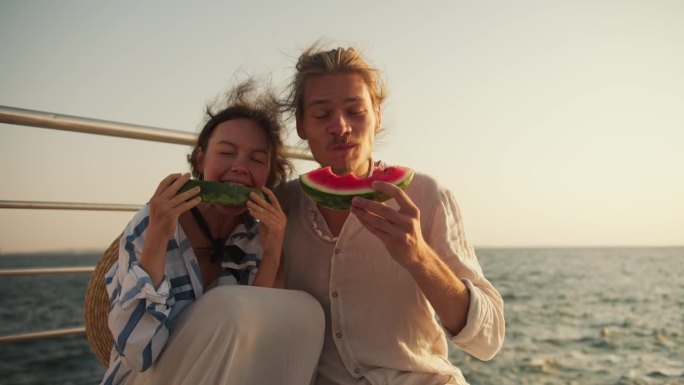 一对幸福的热恋情侣:一个穿着浅色衣服、留着胡茬的金发男孩和一个棕色头发的女孩坐在海边的码头上野餐，吃
