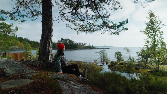 年轻女子独自坐在岩石峭壁上，欣赏美丽的湖景和松林