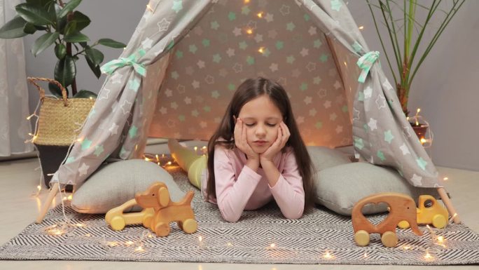 无聊的棕色头发的小女孩在家里客厅的帐篷里玩木制玩具，不开心和无聊，独自一人，没有朋友。