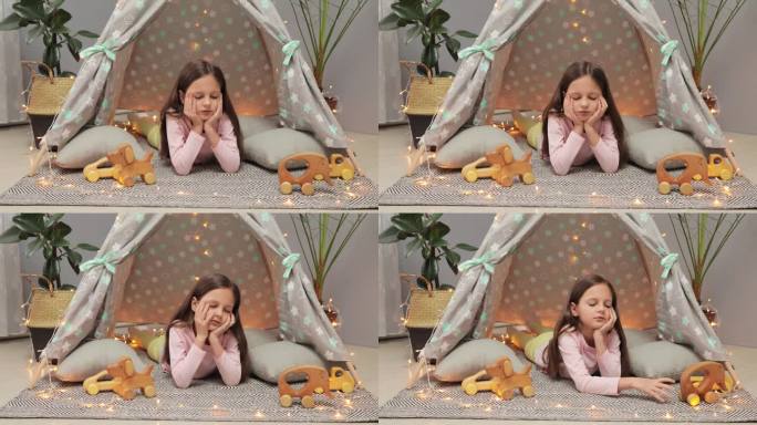 无聊的棕色头发的小女孩在家里客厅的帐篷里玩木制玩具，不开心和无聊，独自一人，没有朋友。