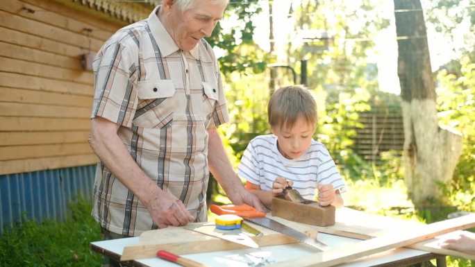 一位白发苍苍的老人在户外教一个男孩做木工活。爷爷教孙子做木匠，用手刨刨木板。