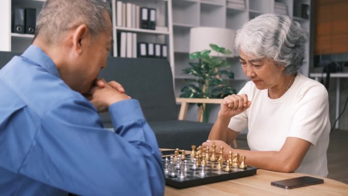 老年人男女穿着休闲装在养老院下棋，围坐在桌子旁，喝茶吃饼干，玩桌上游戏，编织，共度时光