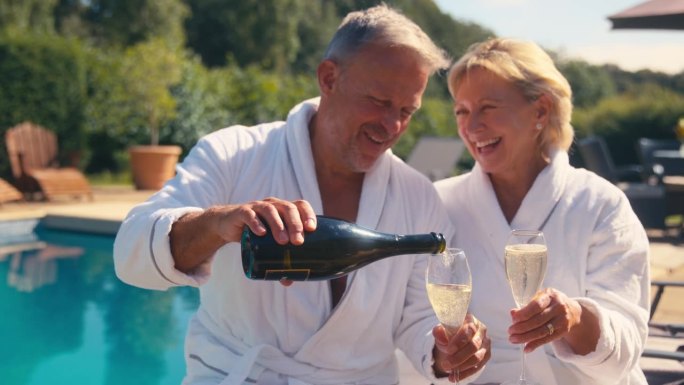 穿着长袍的老夫妇在室外的游泳池边喝香槟