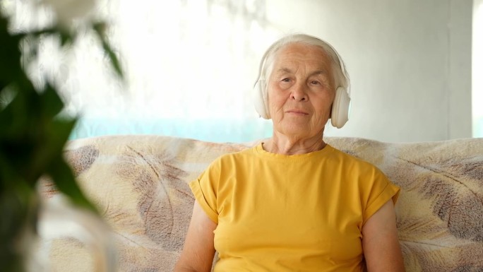戴着耳机的白头发老妇人坐在家里的沙发上，听音乐、有声读物、讲座。概念音频治疗和声音治疗，学习。