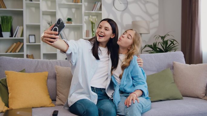 快乐的女孩和妈妈父母在家里用手机拍有趣的照片。一家人微笑着坐在沙发上用智能手机自拍。周末玩得开心放松