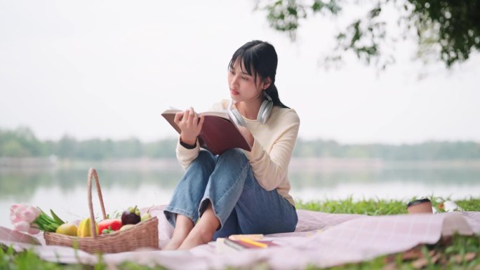 一个亚洲女人在休息日在池塘边野餐，一边看书一边听音乐。一个宁静的湖边时刻，完美的休闲和放松的内容。