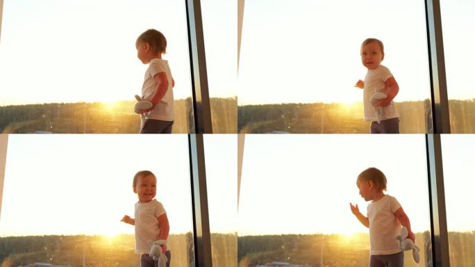 蹒跚学步的小女孩手里拿着玩具，对着窗外的夕阳欣喜不已