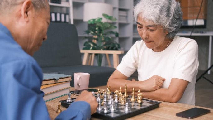 老年人男女穿着休闲装在养老院下棋，围坐在桌子旁，喝茶吃饼干，玩桌上游戏，编织，共度时光