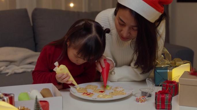 亚洲家庭圣诞快乐，新年快乐。晚上，妈妈和女儿在家里客厅的桌子上烤饼干装饰的糖霜作为圣诞礼物。