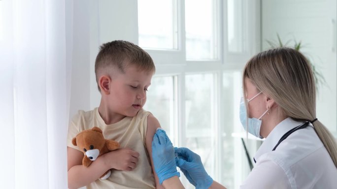 戴着医用口罩的医生给孩子的肩膀打针。疫苗接种,疫苗。护士给孩子注射疫苗。孩子们在诊所里。一个小男孩打